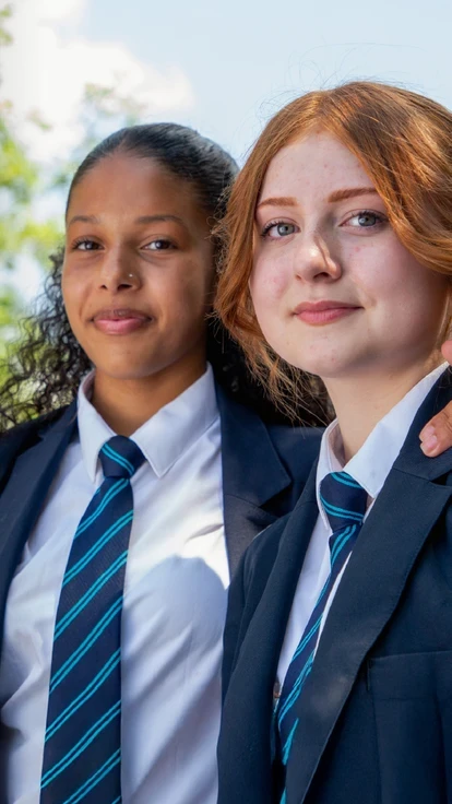 Two secondary school students in uniform smiling together outdoors.