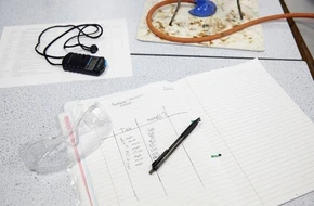 Science experiment setup with safety goggles, stopwatch, and handwritten results in a notebook.