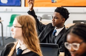 A student in school uniform raises their hand to answer a question in class, while classmates focus on their work.
