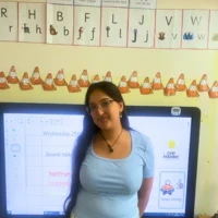 Sanam smiles joyfully in her classroom, stood in front of an interactive whiteboard which is displaying a PowerPoint presentation lesson. Sanam is stood in front of the text, so it is not clear what it says. She is wearing circular glasses with a transparent frame and has long straight black hair. She is wearing a baby blue t-shirt and baby blue and white flowy trousers.